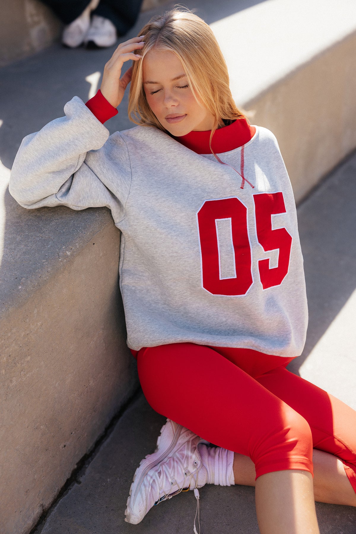 teen, A girl sitting outdoors on concrete steps wearing an oversized light gray sweatshirt with large red '05' lettering, red ribbed cuffs and collar, bright red capris, and white sneakers.
