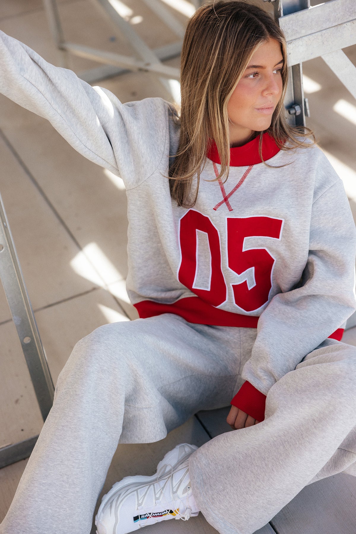 teen, A young girl wearing a light gray sweatshirt and matching sweatpants set with red accents, featuring a large red '05' on the front. She is sitting on a metal bench in a sunlit indoor space.