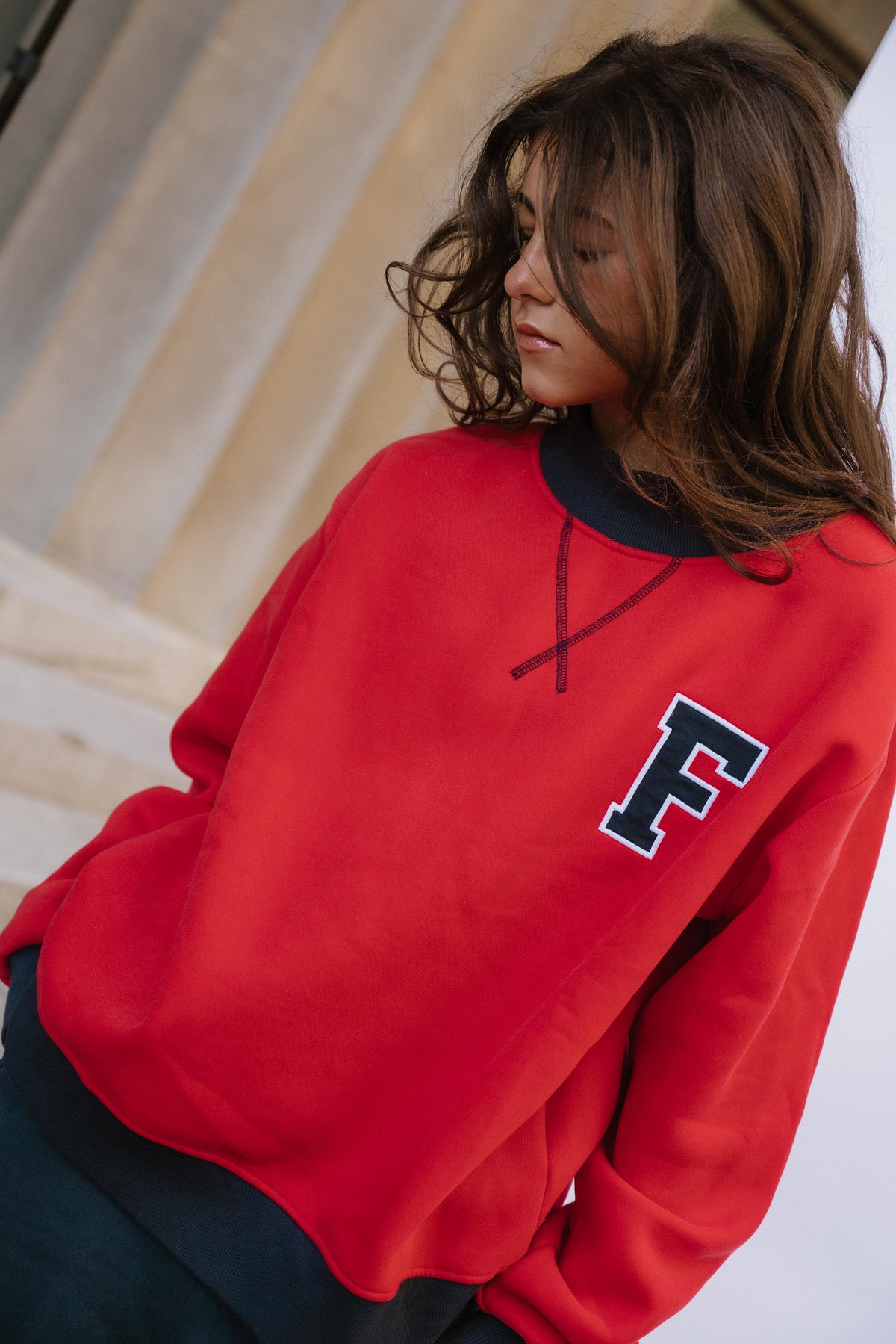 kids, A girl wearing a bright red crewneck sweatshirt with a navy and white block letter 'F' on the left chest, paired with dark pants, set against a blurred beige stone background.