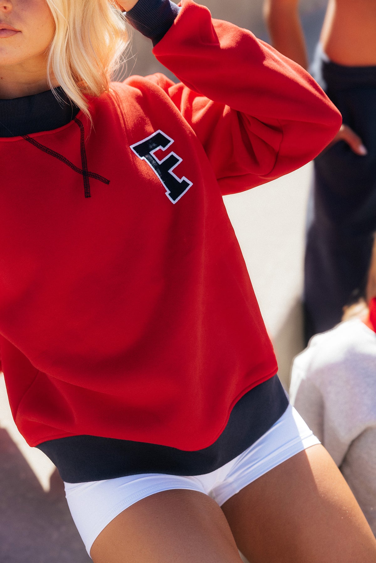 teen, A close-up lifestyle shot of a girl wearing a bright red sweatshirt with navy ribbed trim at the collar, cuffs, and hem, featuring a black and white varsity-style letter 'F' on the left chest. The model is also wearing white shorts.