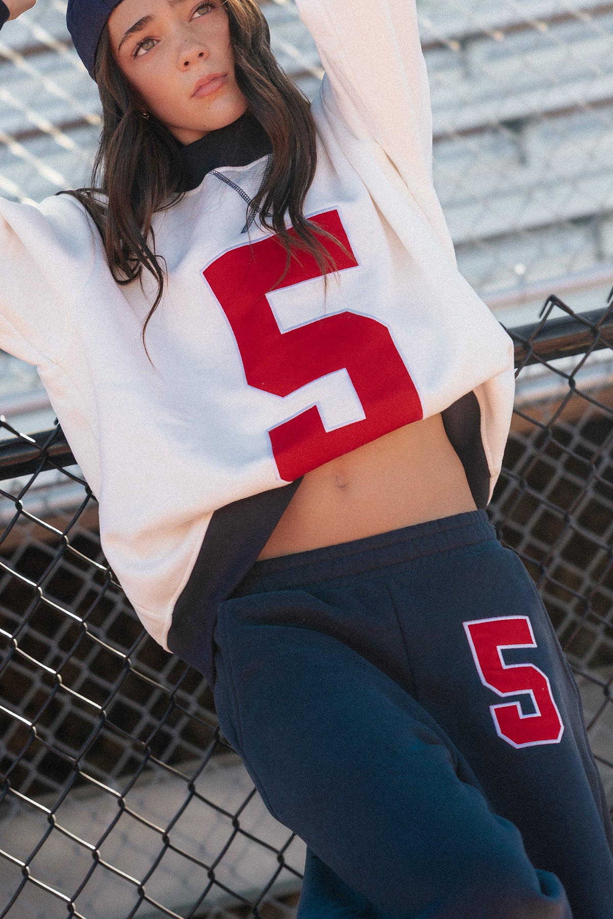 kids, wearing a cream cropped sweatshirt with a large red number 5 on the front, navy sweatpants with a matching red number 5 on the thigh, and a navy baseball cap, leaning against a chain-link fence.
