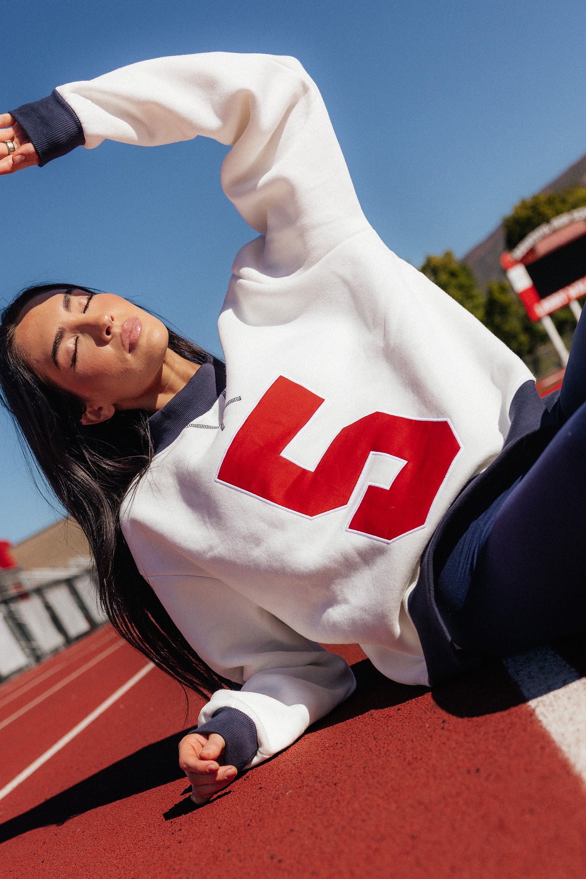 teen, A girl lying on a red running track wearing a white crewneck sweatshirt with a large red number 5 on the front, navy blue pants, and navy blue ribbed cuffs and collar, under a clear blue sky.
