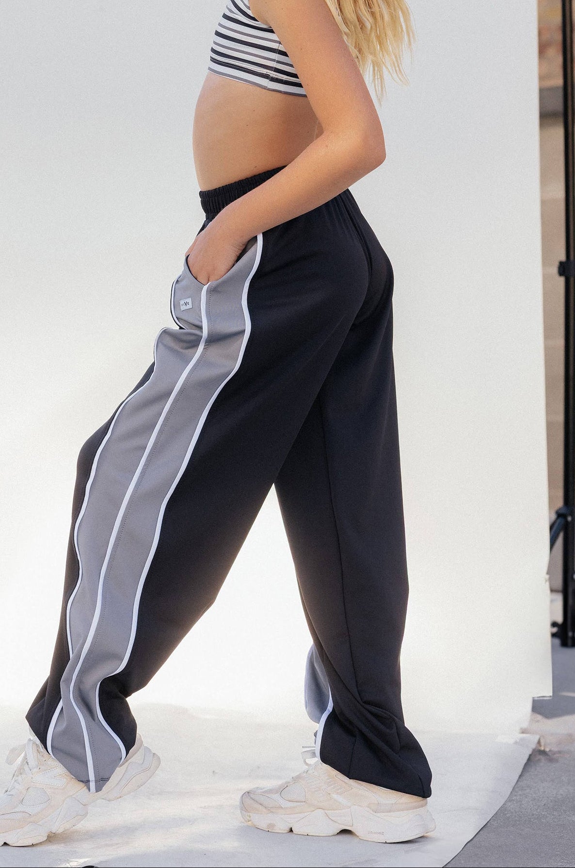 kids, A blonde model wearing black wide-leg track pants with a gray side stripe, a striped crop top, and white sneakers, standing in front of a white studio backdrop.