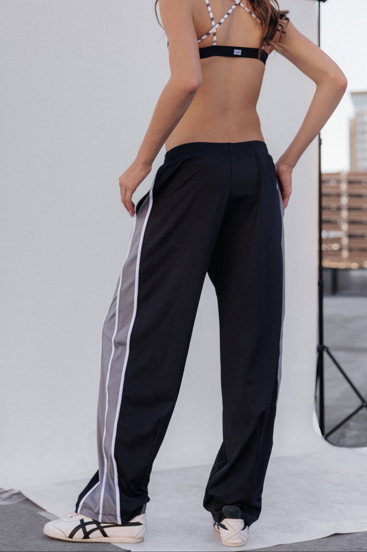 kids,  a girl stands in front of a white studio backdrop wearing loose black track pants with a white side stripe, a black and white strappy bikini top, and white sneakers.