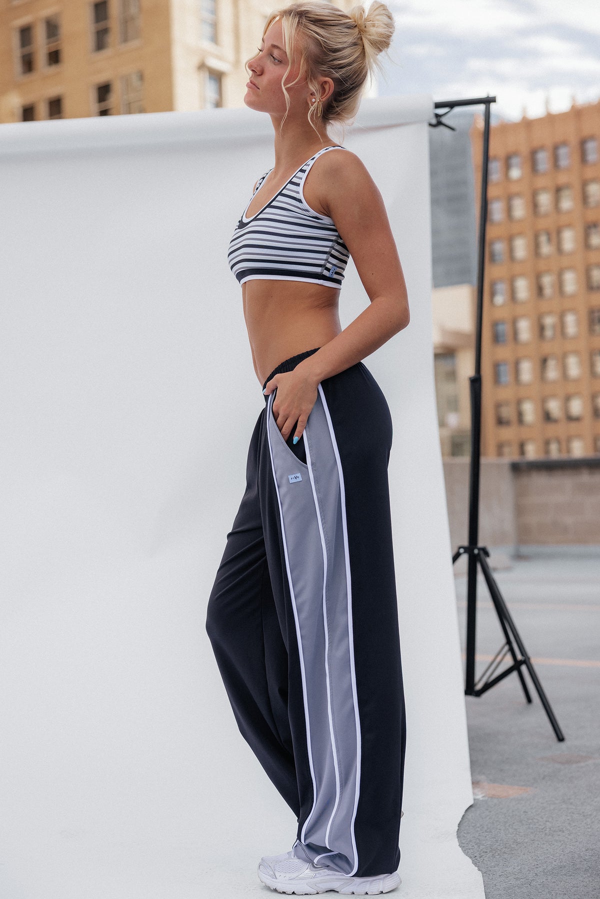 teen, a girl stands in profile wearing a striped sports bra and loose black track pants with a light gray side stripe, posed in front of a white studio backdrop with city buildings in the background.