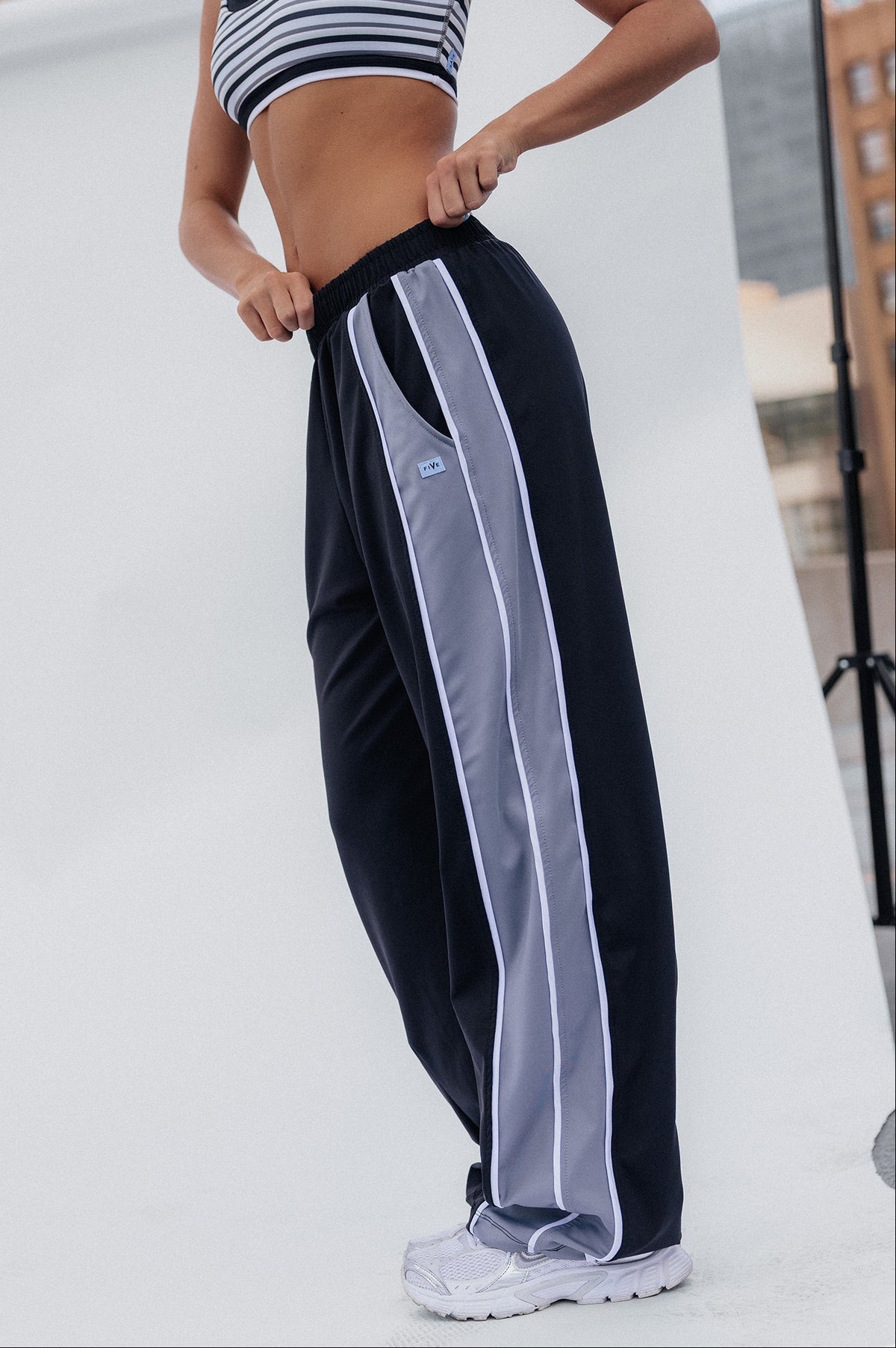 teen, A girl modeling black wide-leg athletic pants with light gray side stripes, paired with a black and white striped sports bra, standing in front of a white studio backdrop with city buildings visible in the background.
