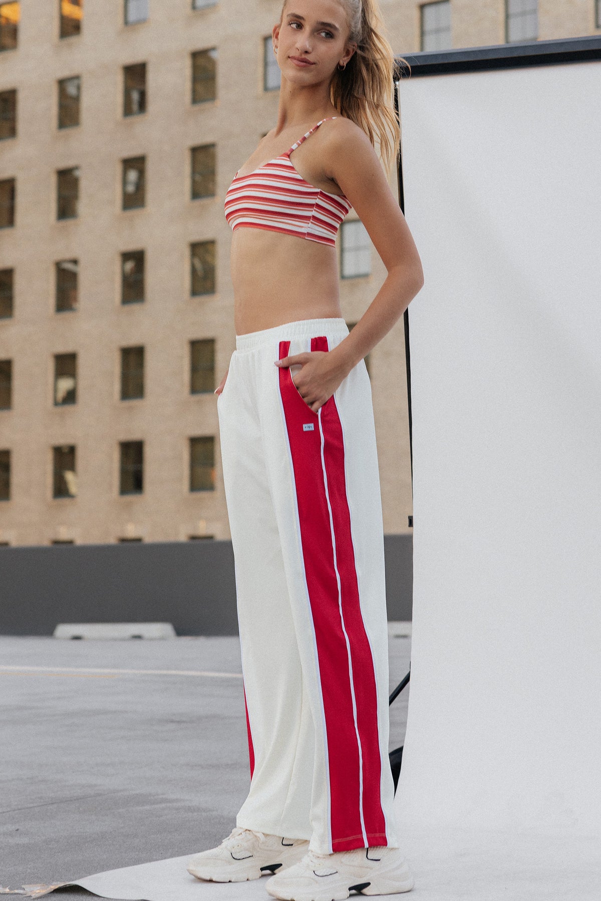 teen, girl stands on a rooftop wearing a red and white striped crop top and white wide-leg sweatpants with bold red side stripes, paired with white sneakers. A large white studio backdrop and a tan building are visible in the background.
