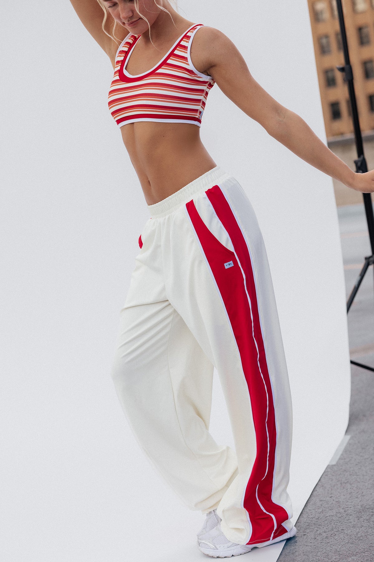 teen, girl modeling a two-piece outfit consisting of a red and white striped crop top and cream track pants with bold red side stripes, photographed in a studio setting.