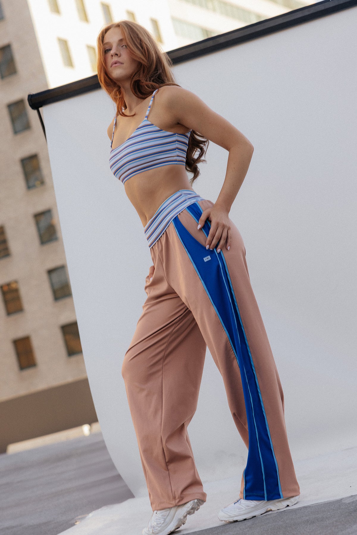 teen, A teen modeling a two-piece activewear set consisting of a blue and white striped crop top and matching wide-leg pants with a blue side stripe, standing in front of a white studio backdrop with a city building in the background.