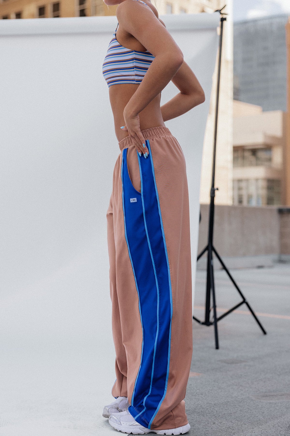teen, girl stands in profile wearing loose peach pants with a bold royal blue side stripe and white sneakers, posed in front of a white studio backdrop with city buildings in the background.