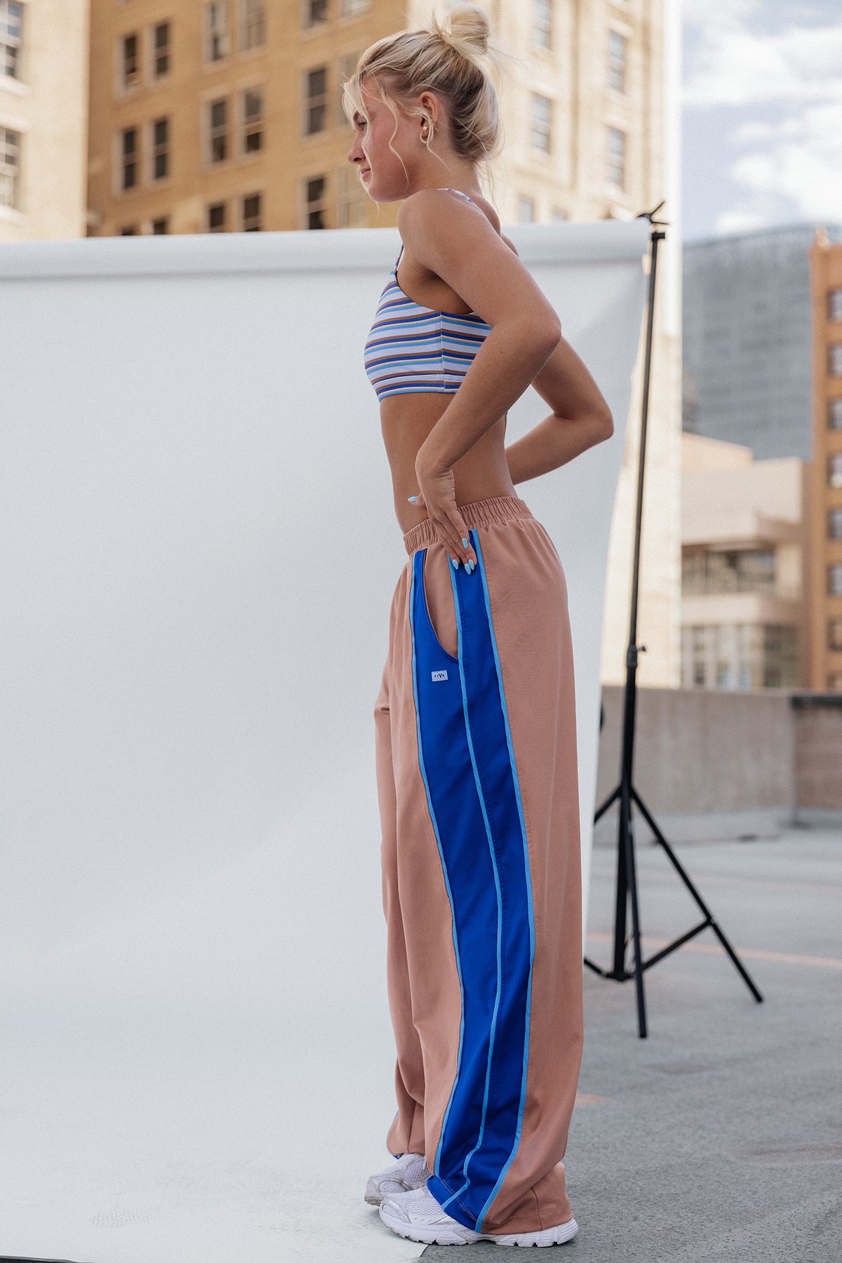 teen, girl stands in profile wearing loose peach pants with a bold royal blue side stripe and white sneakers, posed in front of a white studio backdrop with city buildings in the background.