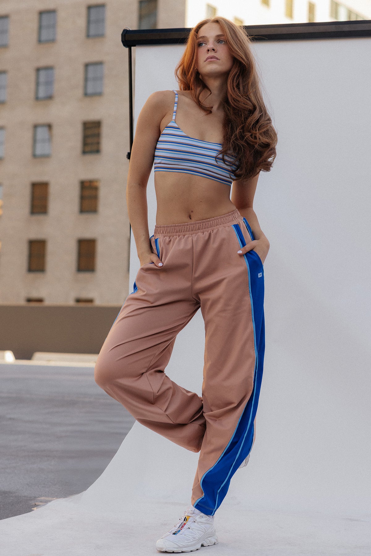 teen, A teen posing outdoors in a matching two-piece activewear set featuring a blue and white striped crop top and beige track pants with blue side stripes.