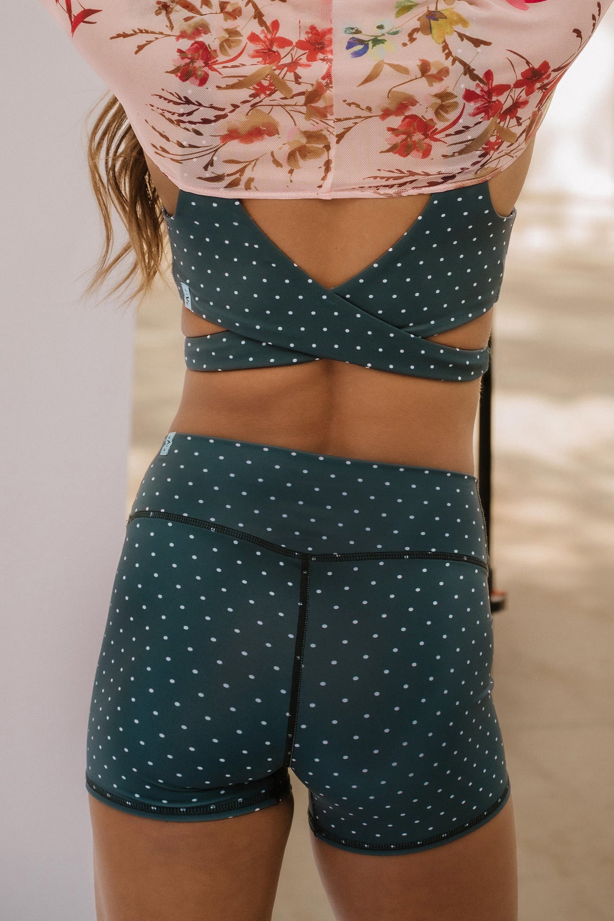 teen, shown from the back wearing a matching dark blue two-piece activewear set with small white polka dots, featuring a crossover back sports bra and high-waisted shorts.