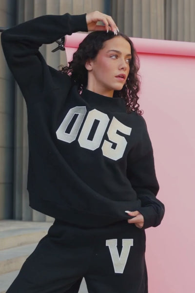 teen, A girl wearing a black sweatshirt with large metallic silver block numbers '005' on the front, posed against a pink wall and stone background.