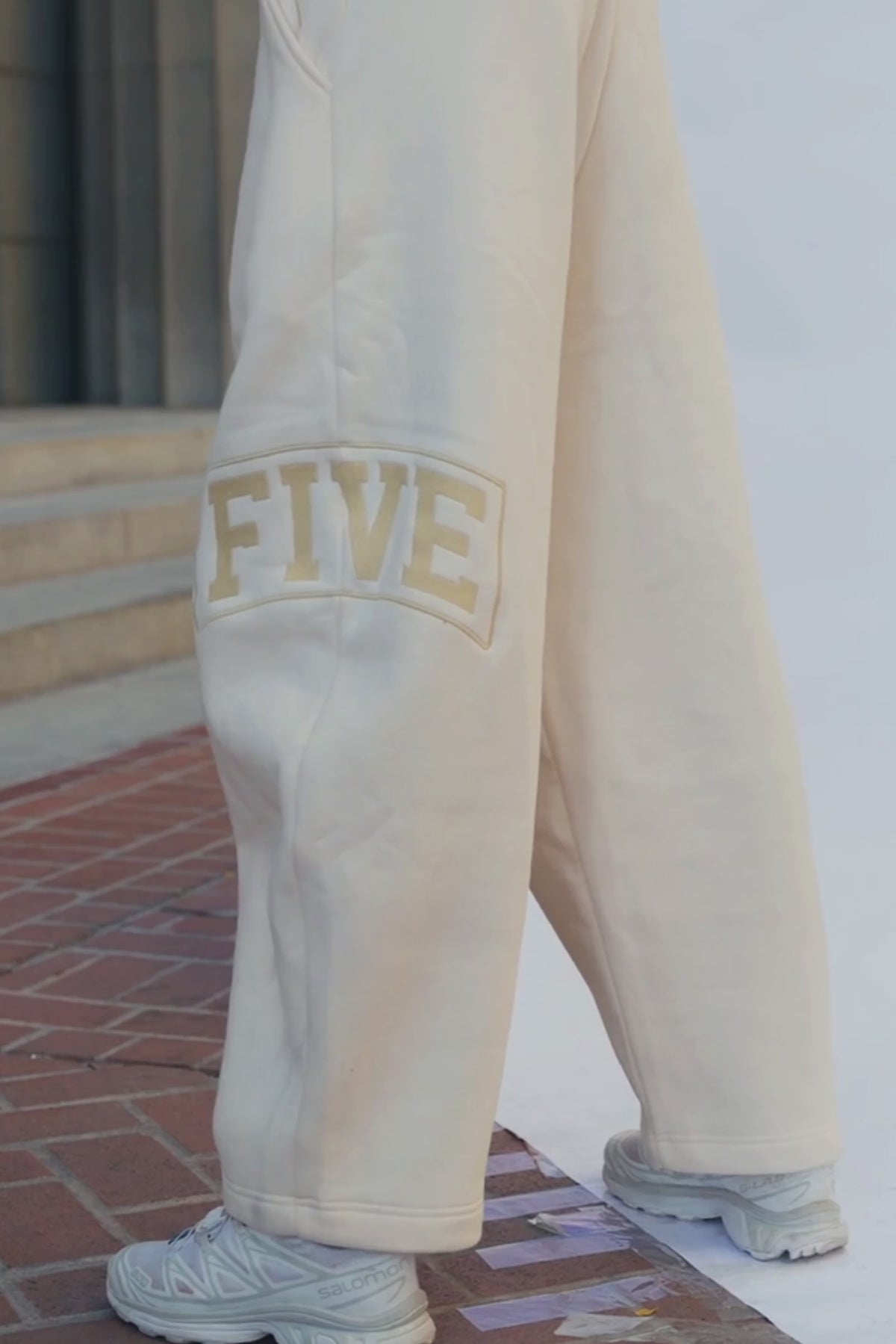 teen, A person wearing cream sweatpants with a gold 'FIVE' graphic on the leg, paired with white sneakers, standing against a pink backdrop.