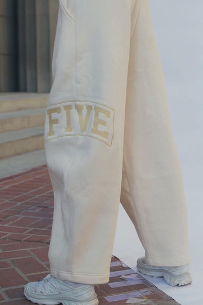 teen, A person wearing cream sweatpants with a gold 'FIVE' graphic on the leg, paired with white sneakers, standing against a pink backdrop.