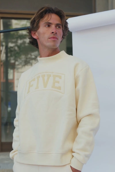 men, A man wearing a cream-colored crewneck sweatshirt with a tonal beige 'FIVE' graphic on the chest, standing on stone steps.
