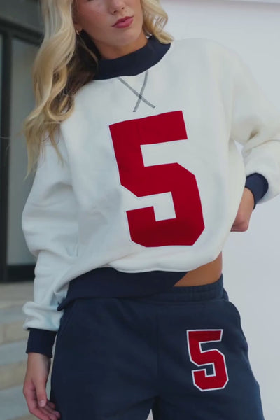 teen, A teen wearing a cream-colored crewneck sweatshirt with a large red number 5 on the front, paired with dark navy sweatpants that also feature a red and white number 5 on the thigh.