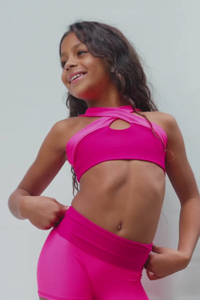 kids, a video of a kid modeling a bright hot pink activewear set consisting of a sports bra with keyhole cutouts and matching high-waisted shorts, posed against a neutral light gray wall.