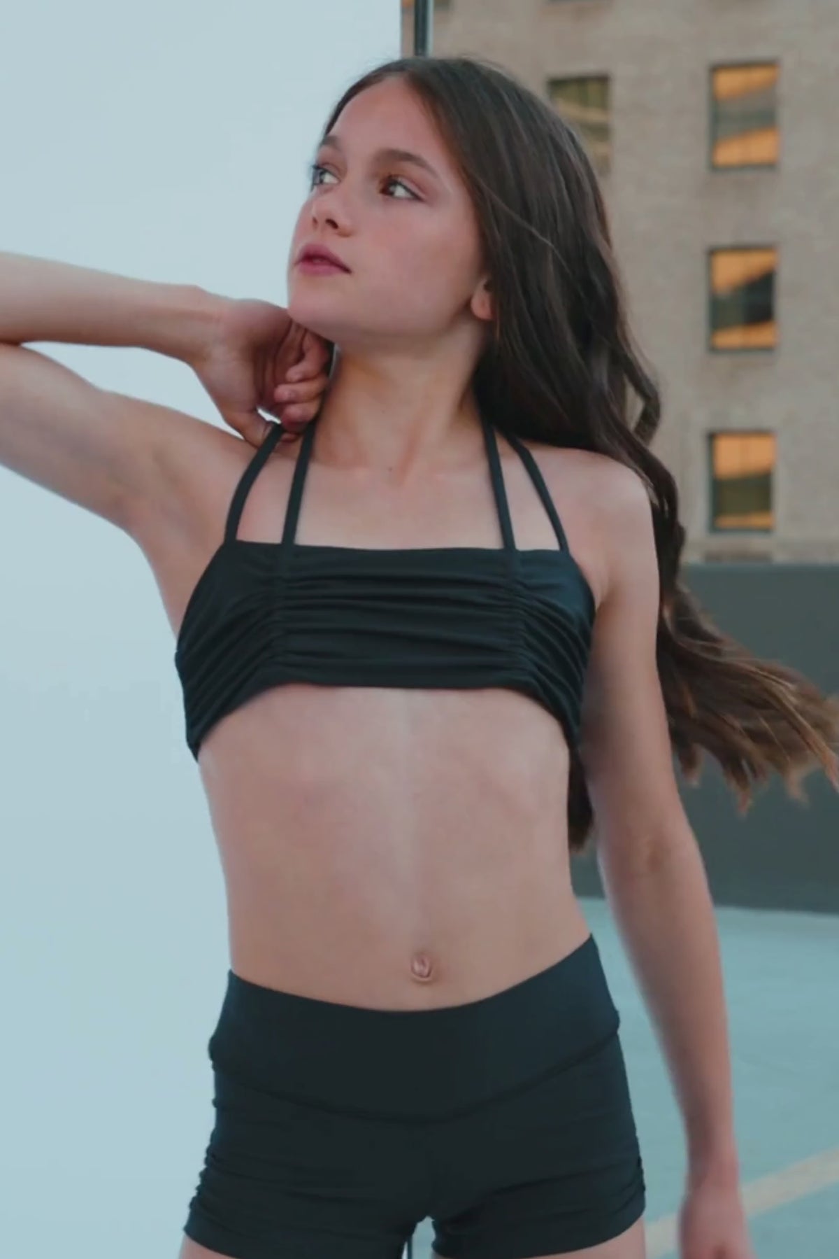 kids, A young woman with dark brown hair poses in a black strappy bra top, looking over her shoulder. The background is a mix of a light gray wall and a darker gray floor, with a diagonal white line dividing the frame.