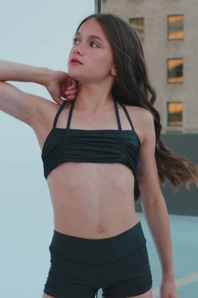 kids, A young woman with dark brown hair poses in a black strappy bra top, looking over her shoulder. The background is a mix of a light gray wall and a darker gray floor, with a diagonal white line dividing the frame.