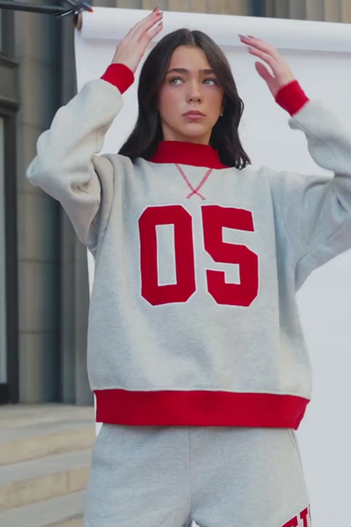 teen, A girl wearing a light heather gray sweatshirt and matching sweatpants set. The sweatshirt has a red crew neck, red ribbed cuffs and hem, and large red '05' lettering outlined in white on the front.