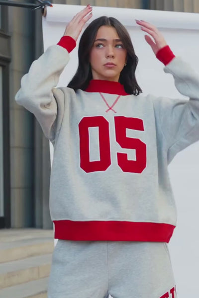teen, A girl wearing a light heather gray sweatshirt and matching sweatpants set. The sweatshirt has a red crew neck, red ribbed cuffs and hem, and large red '05' lettering outlined in white on the front.