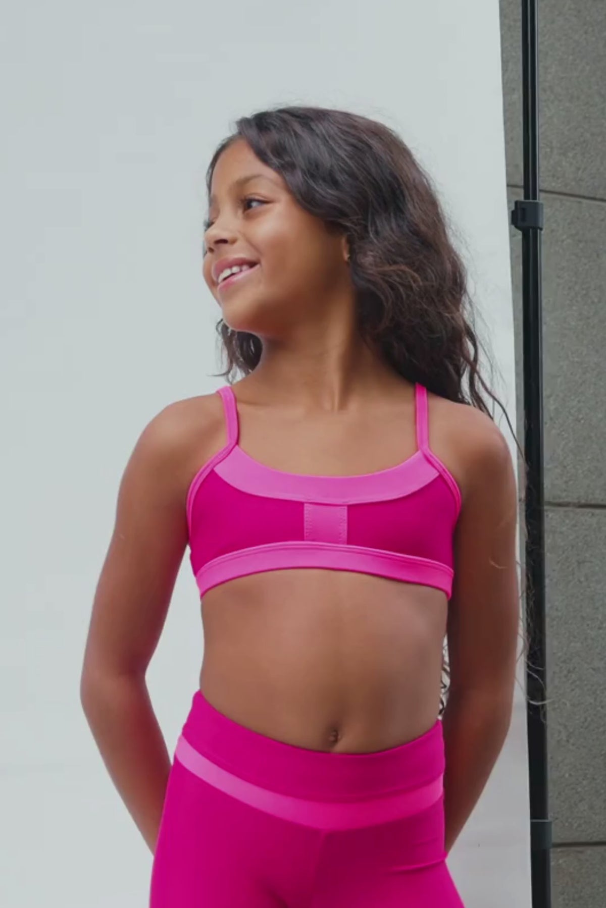 kids, A video of a girl wearing a bright hot pink sports bra and matching high-waisted leggings against a neutral beige background.