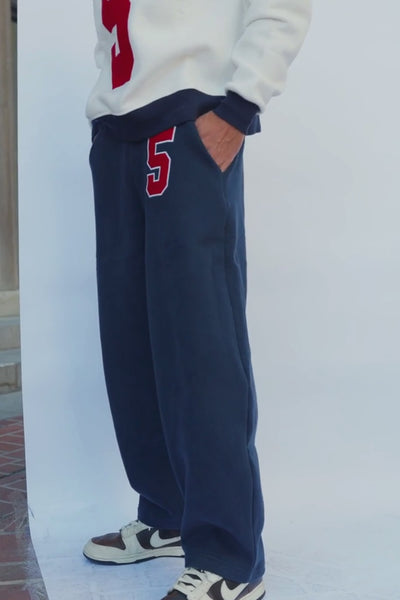 mens, A person wearing dark charcoal gray wide-leg sweatpants with a red and white number 5 on the left thigh, paired with a white sweatshirt and brown and white sneakers.