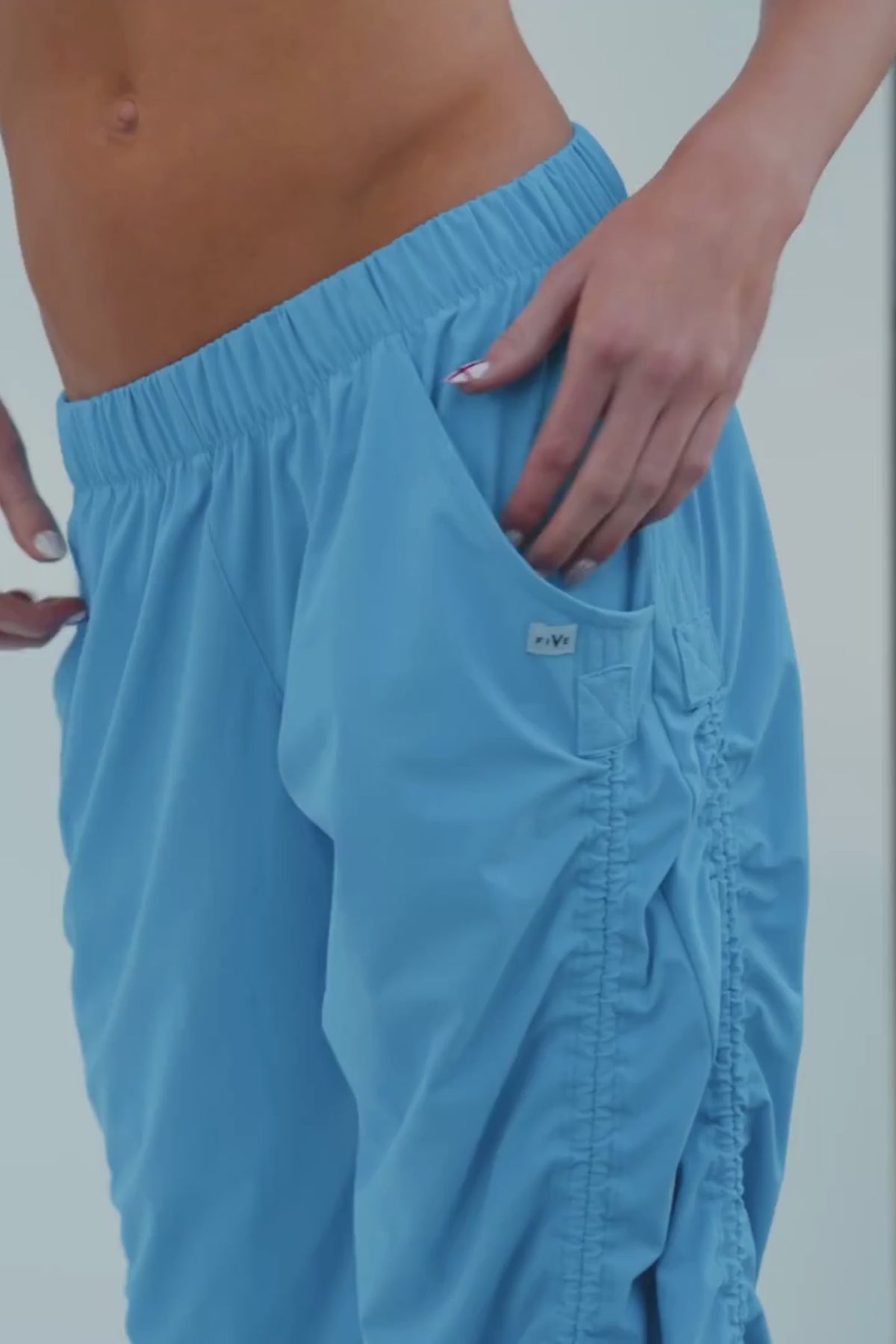 teen, A model wearing bright blue ruched wide-leg sweatpants, styled with a matching blue sports bra and white sneakers, standing on a white platform.