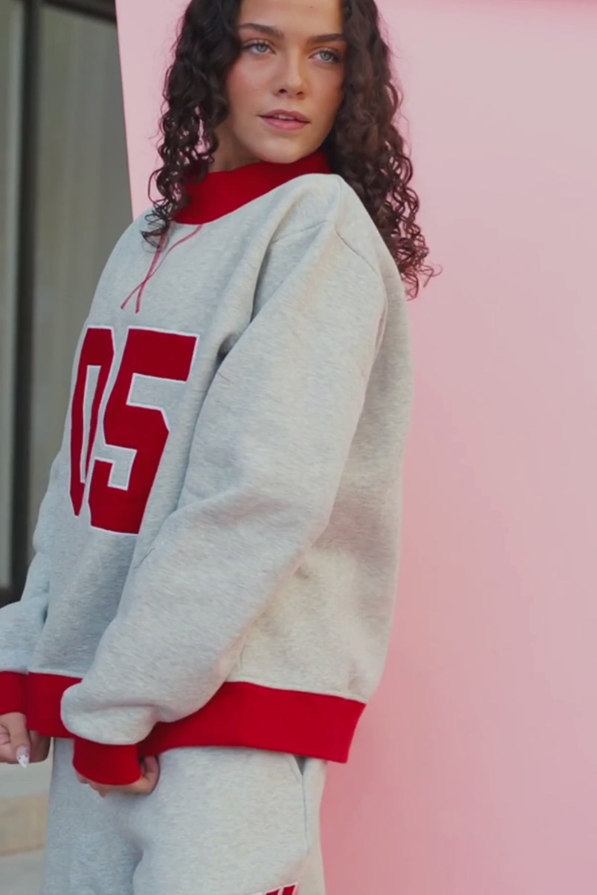 teen, A girl wearing a light heather gray sweatshirt and matching sweatpants set. The sweatshirt has a red crew neck, red ribbed cuffs and hem, and large red '05' lettering outlined in white on the front.