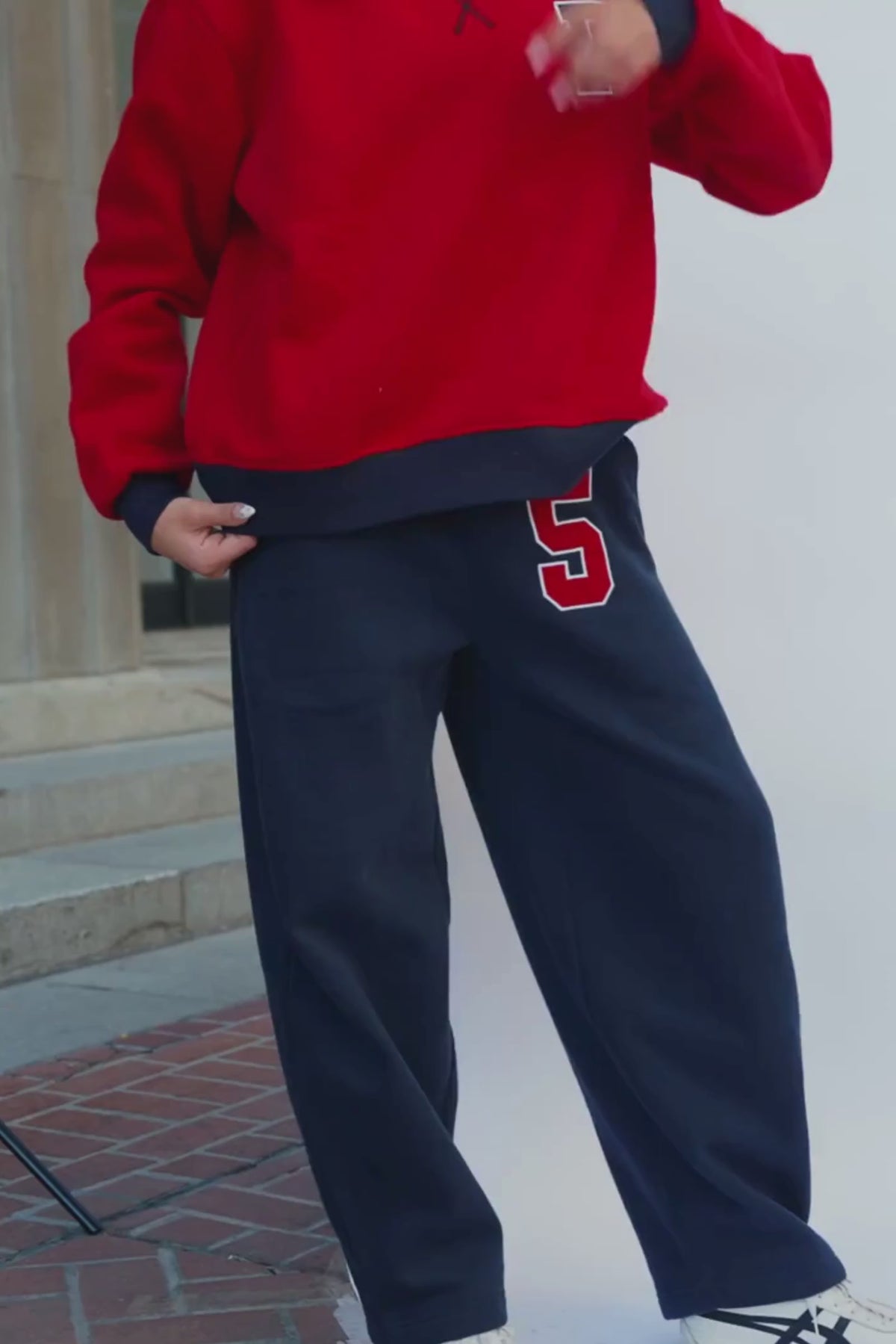 teen, modeling a pair of navy sweatpants with a red and white number 5 on the left thigh, paired with a red sports bra, standing against a white studio backdrop.