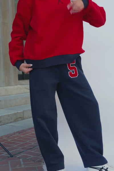 teen, modeling a pair of navy sweatpants with a red and white number 5 on the left thigh, paired with a red sports bra, standing against a white studio backdrop.