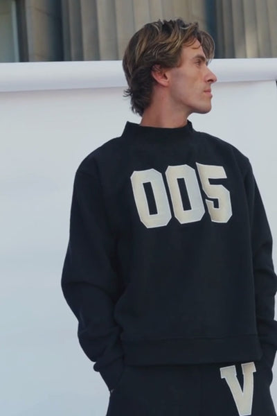 mens, A man wearing a black sweatshirt with large white '005' lettering on the front and matching black pants with a white 'V' graphic, photographed in an indoor setting with steps in the background.