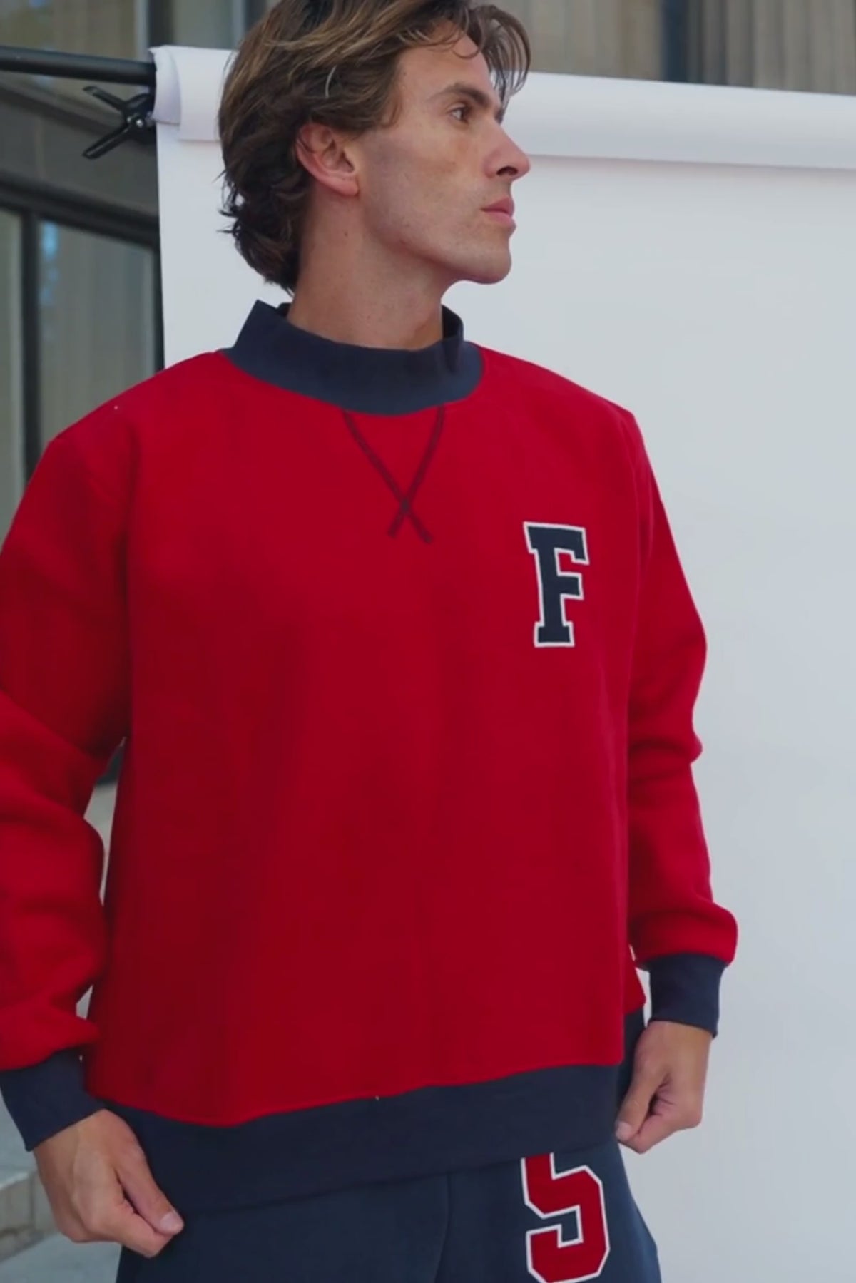 mens, A man wearing a bright red crewneck sweatshirt with a dark navy collar, cuffs, and hem, featuring a large navy block letter 'F' outlined in white on the left chest. He is looking down, with his hands in his pockets, wearing dark navy sweatpants.
