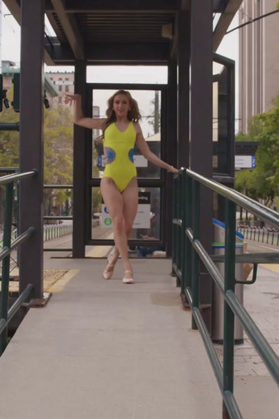 teen, a dancer dancing for a video against a street sign in a bright yellow leotard with turquoise starburst cutouts.