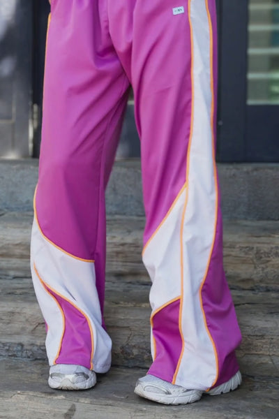 teen, A person wearing wide-leg athletic pants in a bright pink-purple color with a white side stripe bordered by orange piping, standing against a pale pink wall with a strong shadow