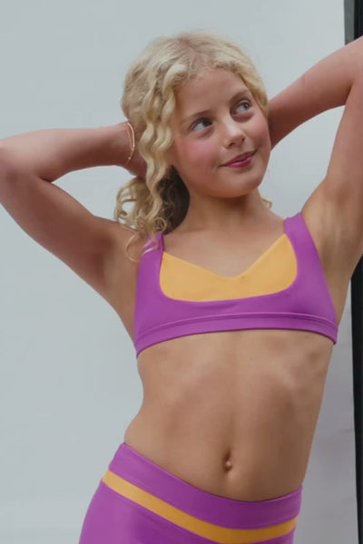 kids, A video of a young girl wearing a two-tone athletic sports bra and matching high-waisted leggings in purple and orange, with her blonde curly hair visible.