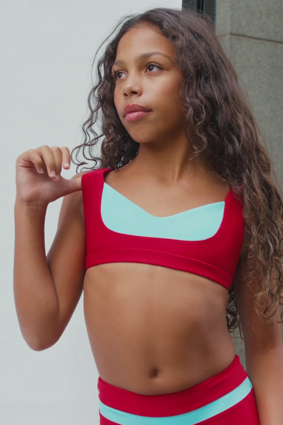 kids, kid wearing a matching red and light blue athletic set, featuring a sports bra and high-waisted leggings, posing against a neutral concrete wall.