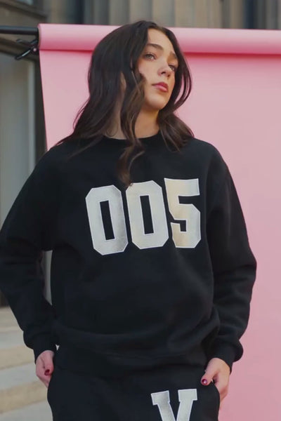 kids, A girl wearing a black sweatshirt with large silver varsity-style '005' lettering on the front and matching black sweatpants with a large white 'V' on the thigh, posed against a pink wall.
