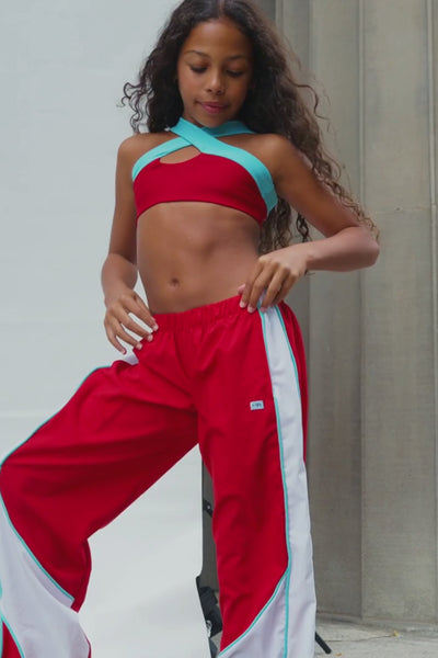 kids, a video of a kid wearing red and white athletic track pants with a light blue side stripe, paired with a matching red sports bra and white sneakers, posing on outdoor stone steps.