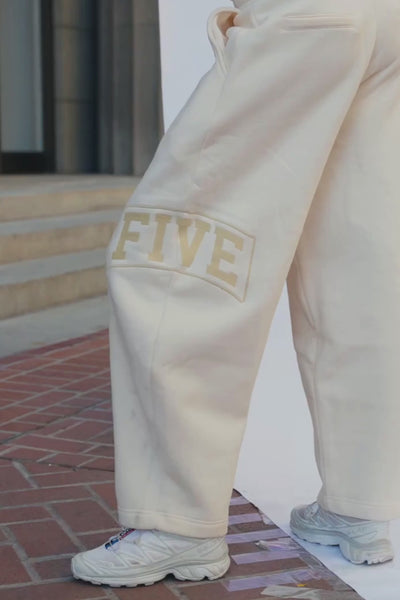 kids, A girl wearing loose, wide-leg butter yellow sweatpants with side pockets and a large yellow-toned logo embroidered on the lower left leg, paired with white sneakers, standing outdoors on a brick and concrete surface.