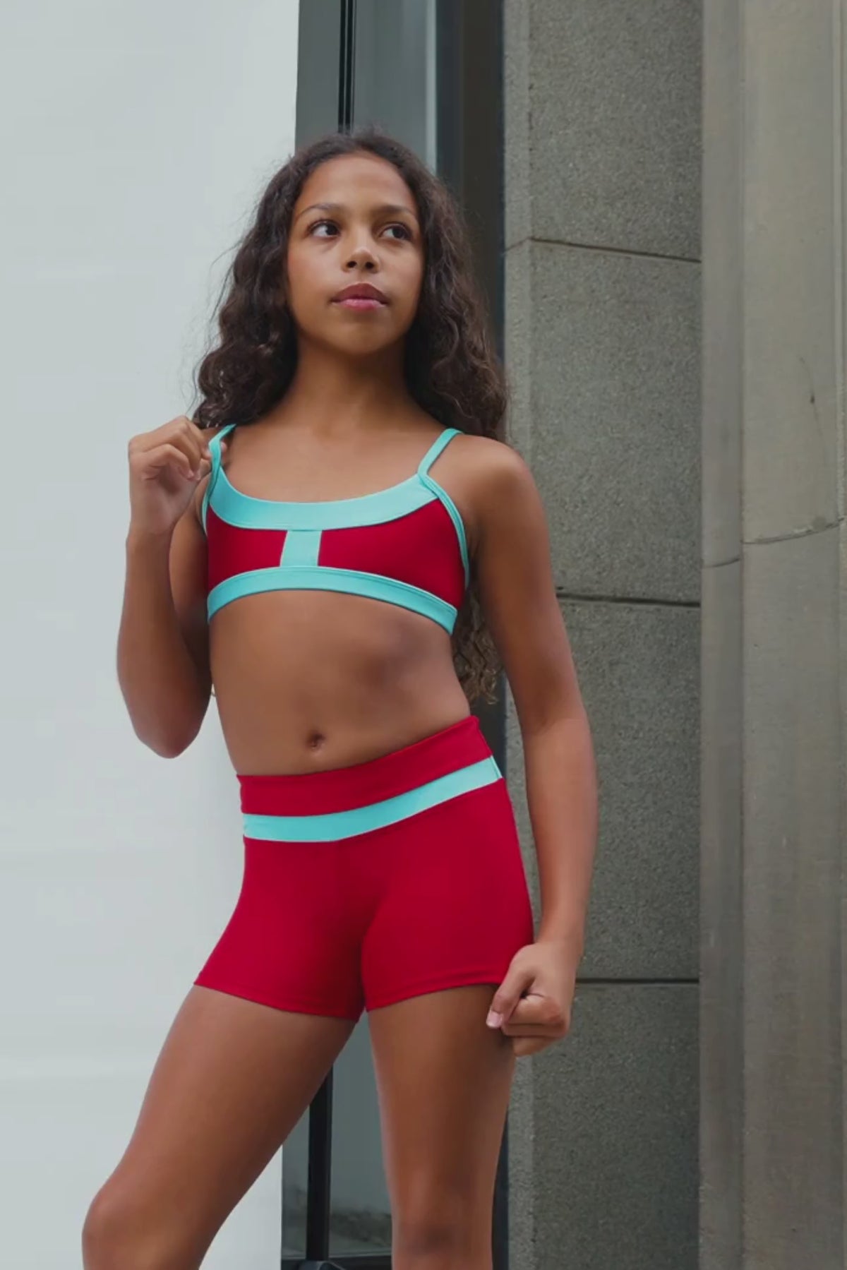 kids, A model wearing a matching red and turquoise athletic set consisting of a sports bra and high-waisted shorts, posing against a neutral concrete wall.