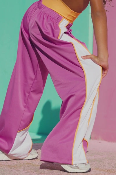 kids,  young girl posing in a vibrant purple and orange athletic crop top with matching purple sweatpants featuring white side panels and orange piping, set against a colorful teal and pink wall.