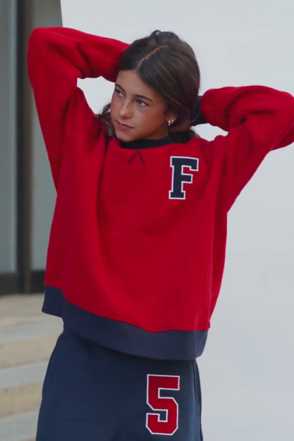 kids, A girl wearing a bright red crewneck sweatshirt with a navy ribbed collar, cuffs, and hem, featuring a navy collegiate letter 'F' on the left chest. She is also wearing navy sweats with a red and white '5' on the thigh.
