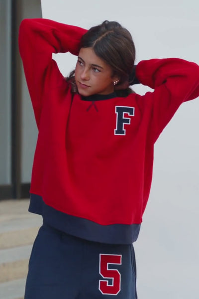 kids, A girl wearing a bright red crewneck sweatshirt with a navy ribbed collar, cuffs, and hem, featuring a navy collegiate letter 'F' on the left chest. She is also wearing navy sweats with a red and white '5' on the thigh.