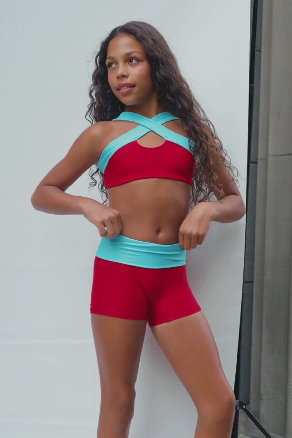 kids, A kid modeling a matching red activewear set with light blue trim, consisting of a sports bra and shorts, posed on a white studio backdrop next to a concrete wall.