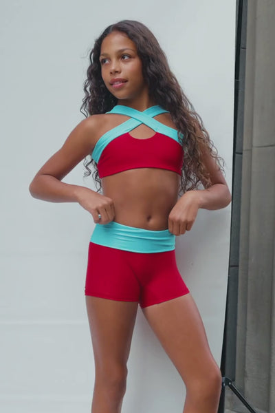kids, A kid modeling a matching red activewear set with light blue trim, consisting of a sports bra and shorts, posed on a white studio backdrop next to a concrete wall.