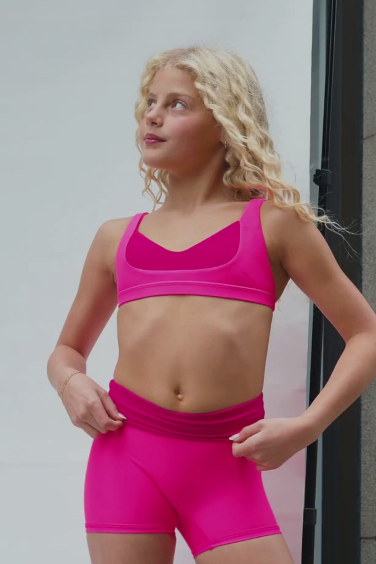 kids, A video of a young girl with curly blonde hair wearing a bright hot pink sports bra and matching pink bottoms, looking off-camera against a neutral background.