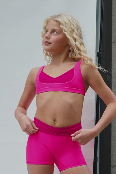 kids, A video of a young girl with curly blonde hair wearing a bright hot pink sports bra and matching pink bottoms, looking off-camera against a neutral background.